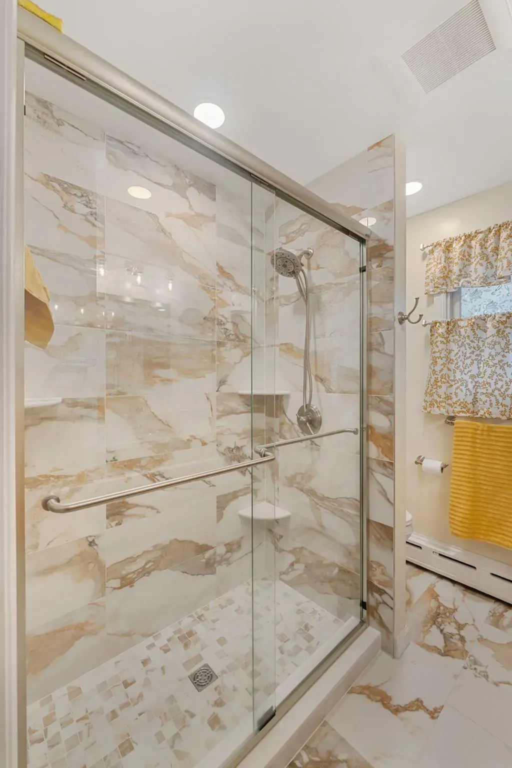 Modern bathroom shower with glass sliding door, beige marble-patterned tile, and a yellow towel rail In Bucks County PA