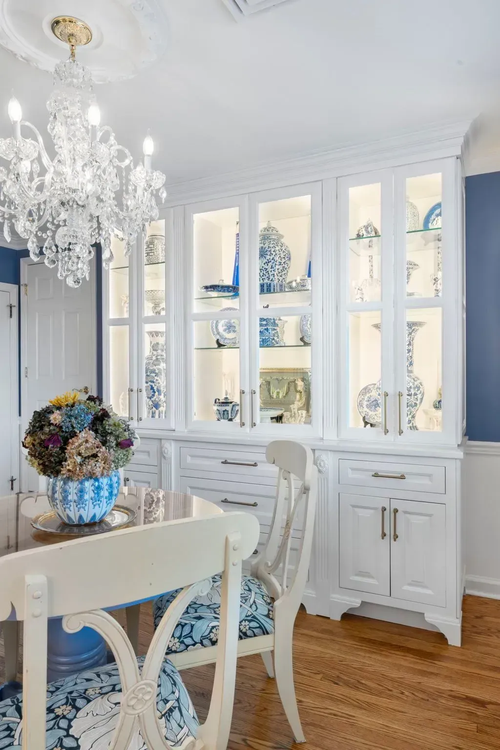 Elegant dining room with crystal chandelier, white glass-front cabinets, blue floral chairs, and wooden floor  In Bucks County PA