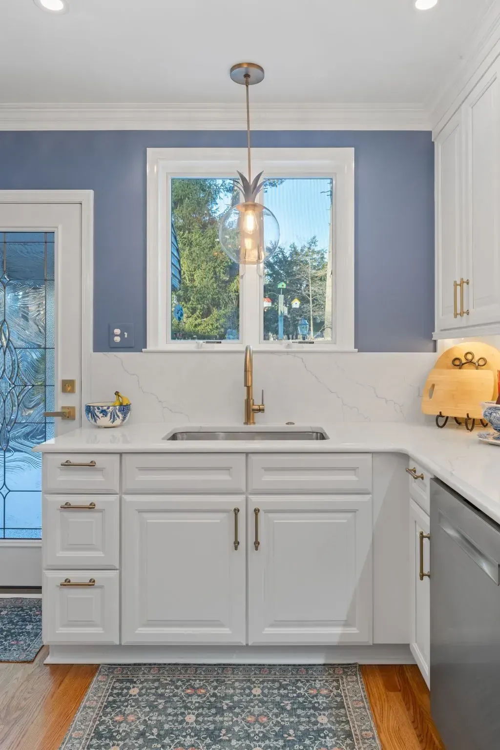 Bright kitchen sink nook with white cabinets, blue walls, gold faucet, and pendant light by a window  In Bucks County PA