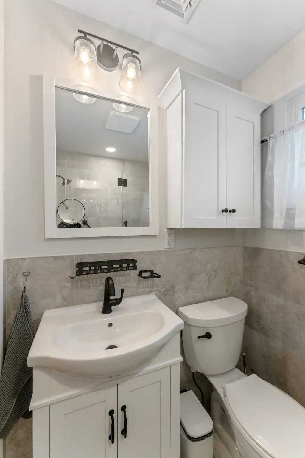 Small bathroom with white vanity, mirror cabinet, toilet, and shower curtain on gray tiled walls  In Bucks County PA
