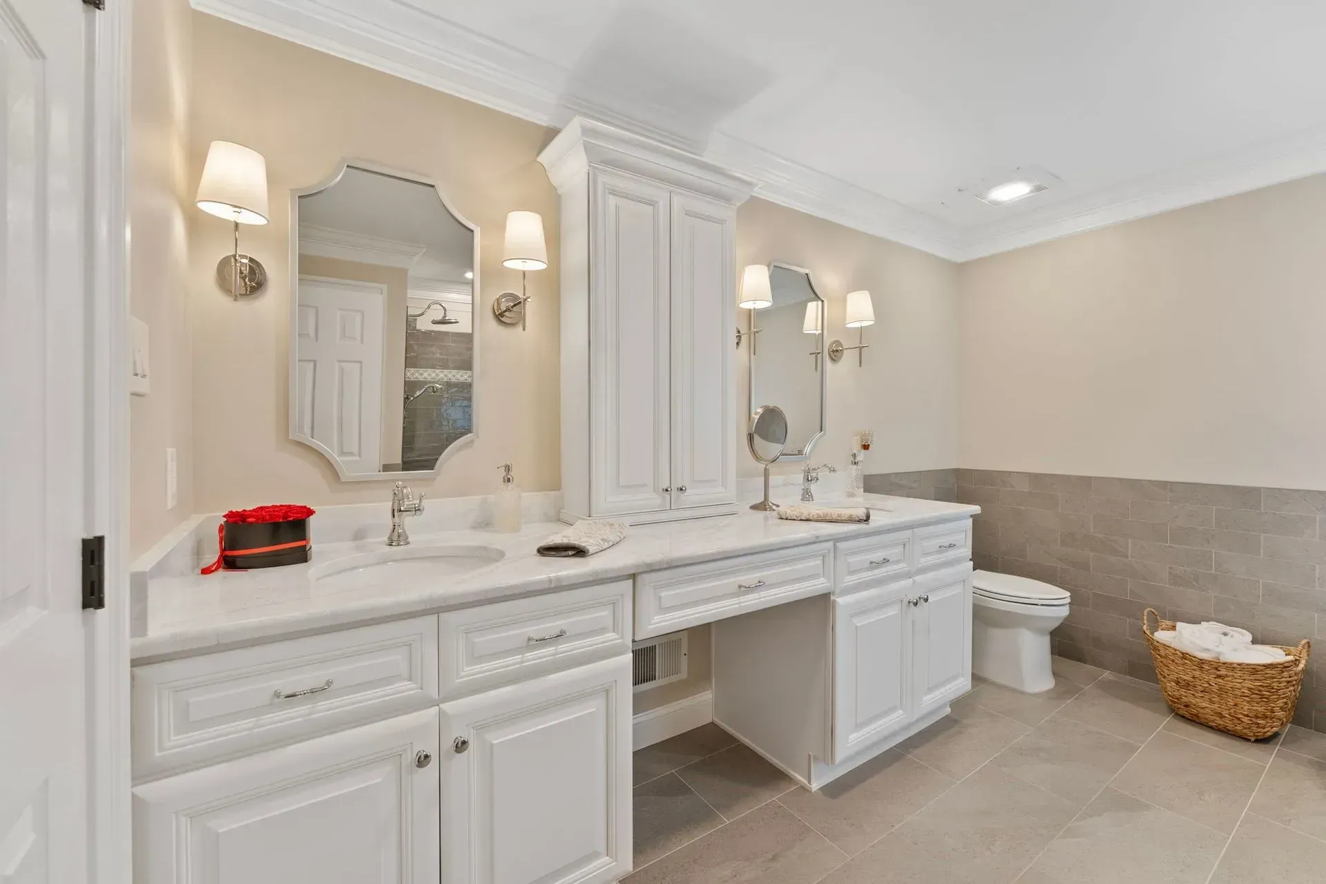 Bright bathroom with white double vanity, large mirrors, beige walls, tiled floor, and toilet with basket nearby In Bucks County PA