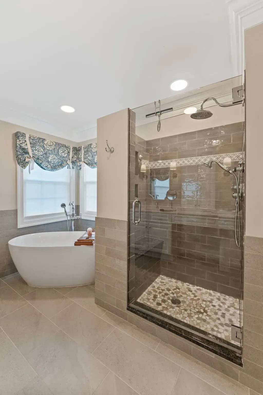 Bright bathroom with a freestanding tub, glass shower, beige tile, and window with patterned drapes In Bucks County PA
