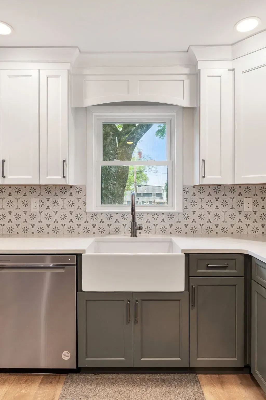 White kitchen with farmhouse sink, gray lower cabinets, stainless dishwasher, and window over patterned backsplash In Bucks County PA