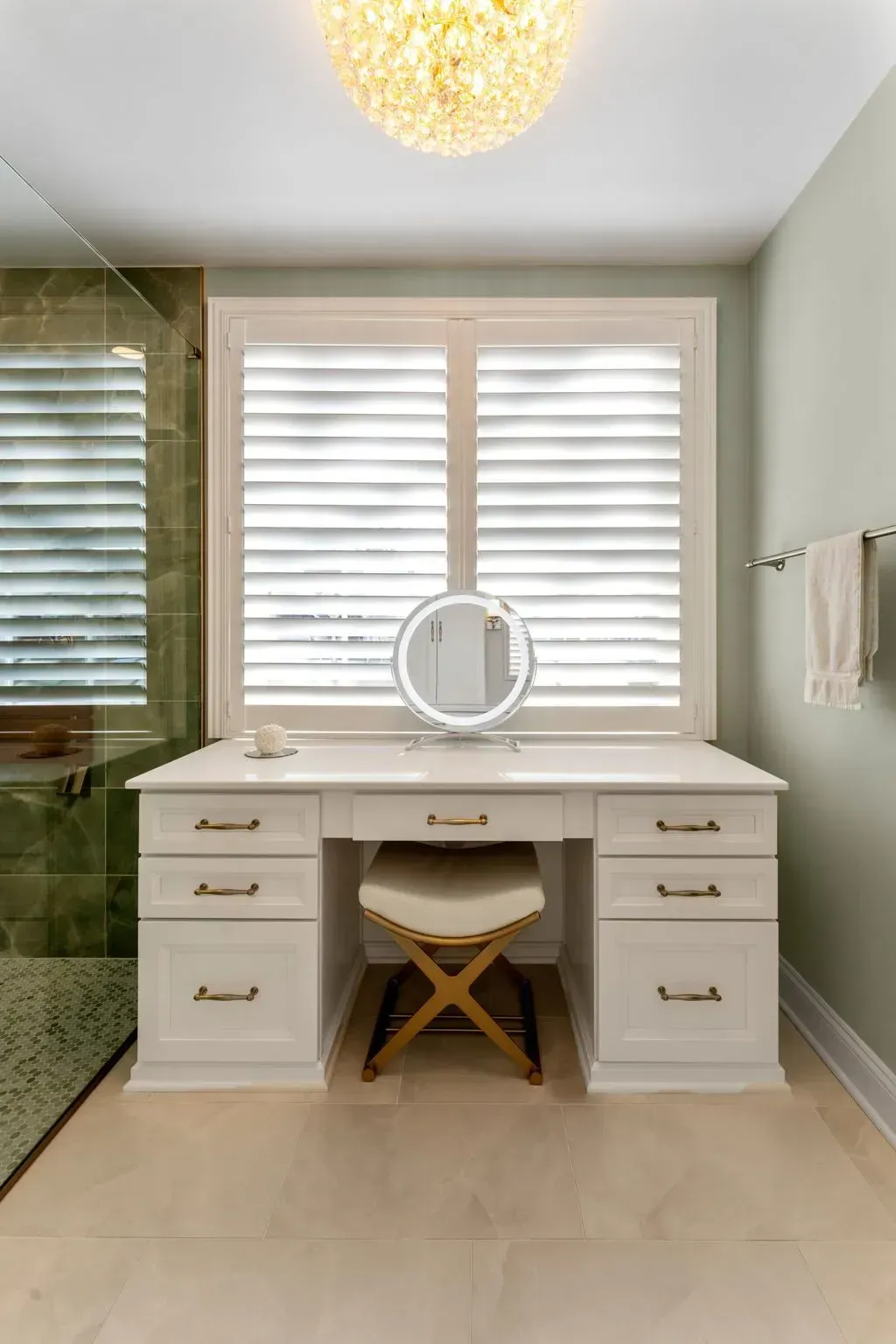 White vanity desk with round mirror in a bright bathroom, flanked by drawers and a stool under window blinds In Bucks County PA