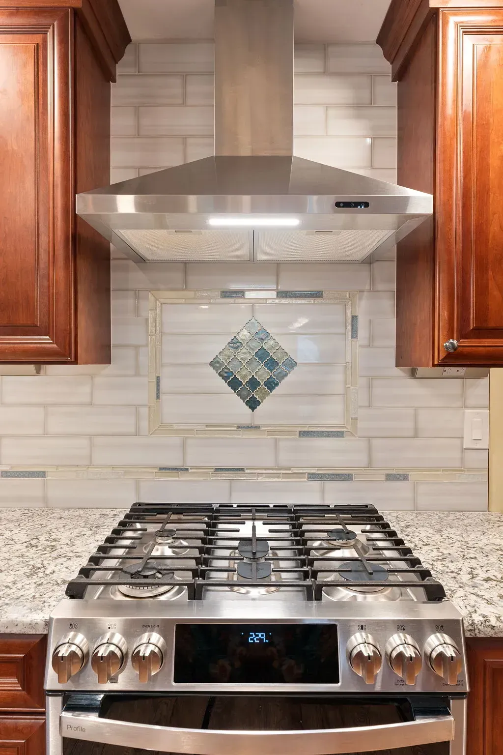 Modern kitchen stove with stainless steel range hood, wood cabinets, and tiled backsplash In Bucks Country PA