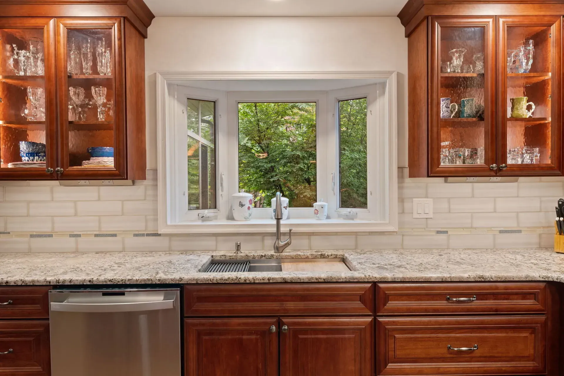 Kitchen with wooden cabinets, granite counters, stainless dishwasher, and a window over the sink overlooking greenery In Bucks Country PA