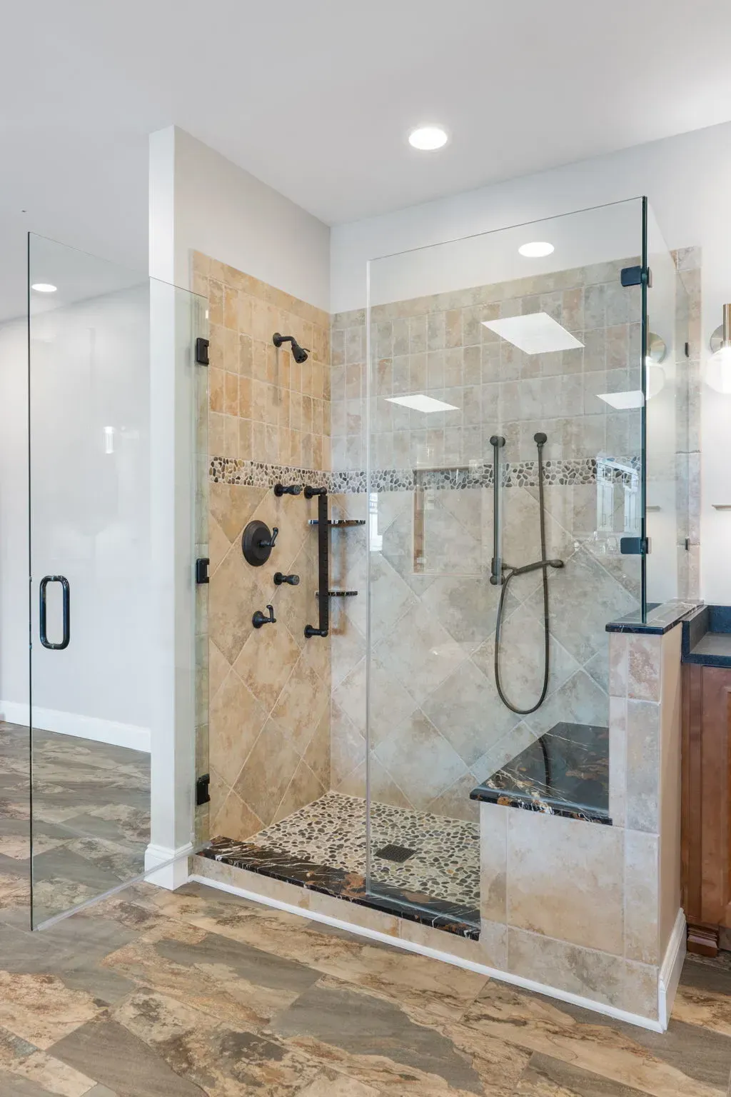 Modern glass walk-in shower with beige tile, black fixtures, and pebble floor in a bright bathroom  In Bucks County PA