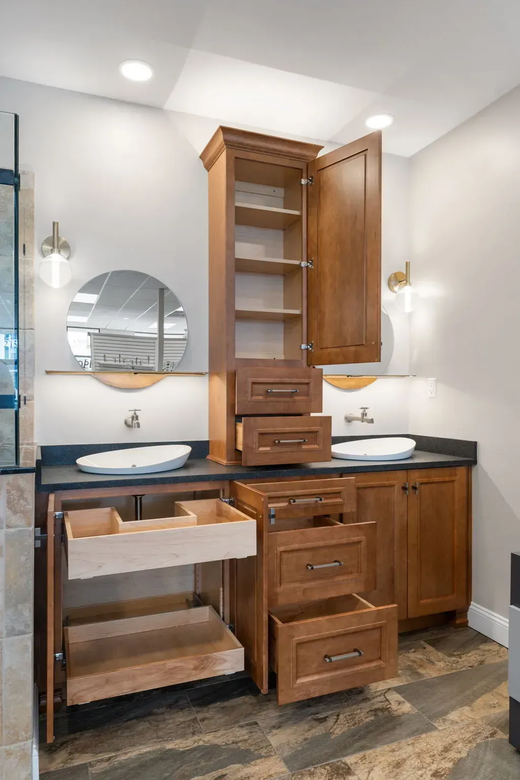 Bathroom vanity with open wood cabinetry, black countertop, round mirror, and white sinks  In Bucks County PA