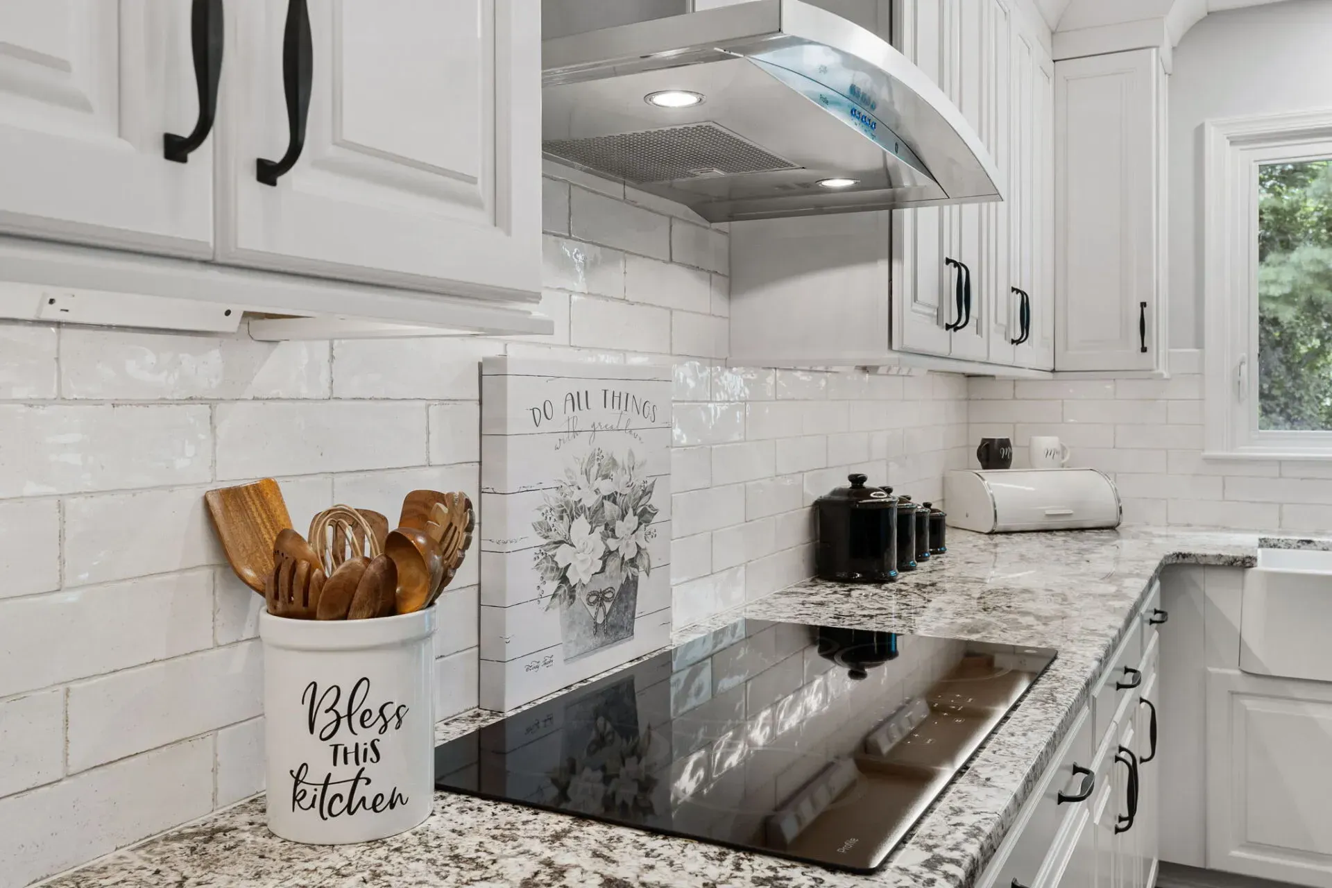 Bright white kitchen with stainless range hood, granite counters, and black cooktop beside window and utensil jar In Bucks County PA