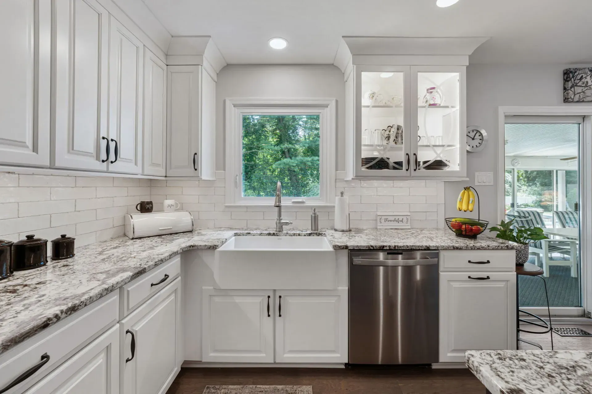 Bright white kitchen with granite counters, farmhouse sink, stainless dishwasher, and window view of greenery In Bucks County PA