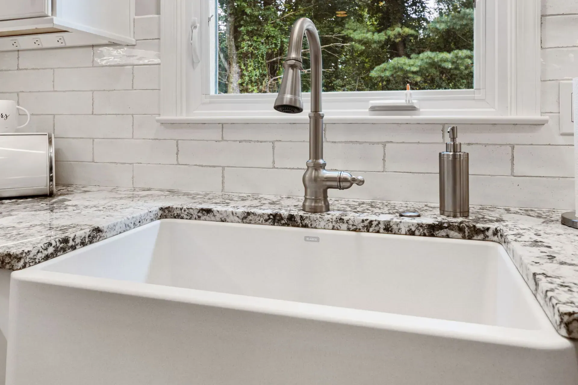 Modern kitchen sink with white farmhouse basin, chrome faucet, and window above granite countertop In Bucks County PA