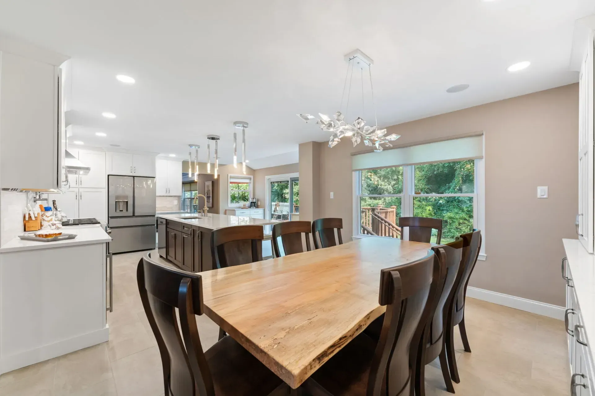 Bright modern dining area with a large wooden table, dark chairs, kitchen island, and sliding glass door to greenery In Bucks County PA