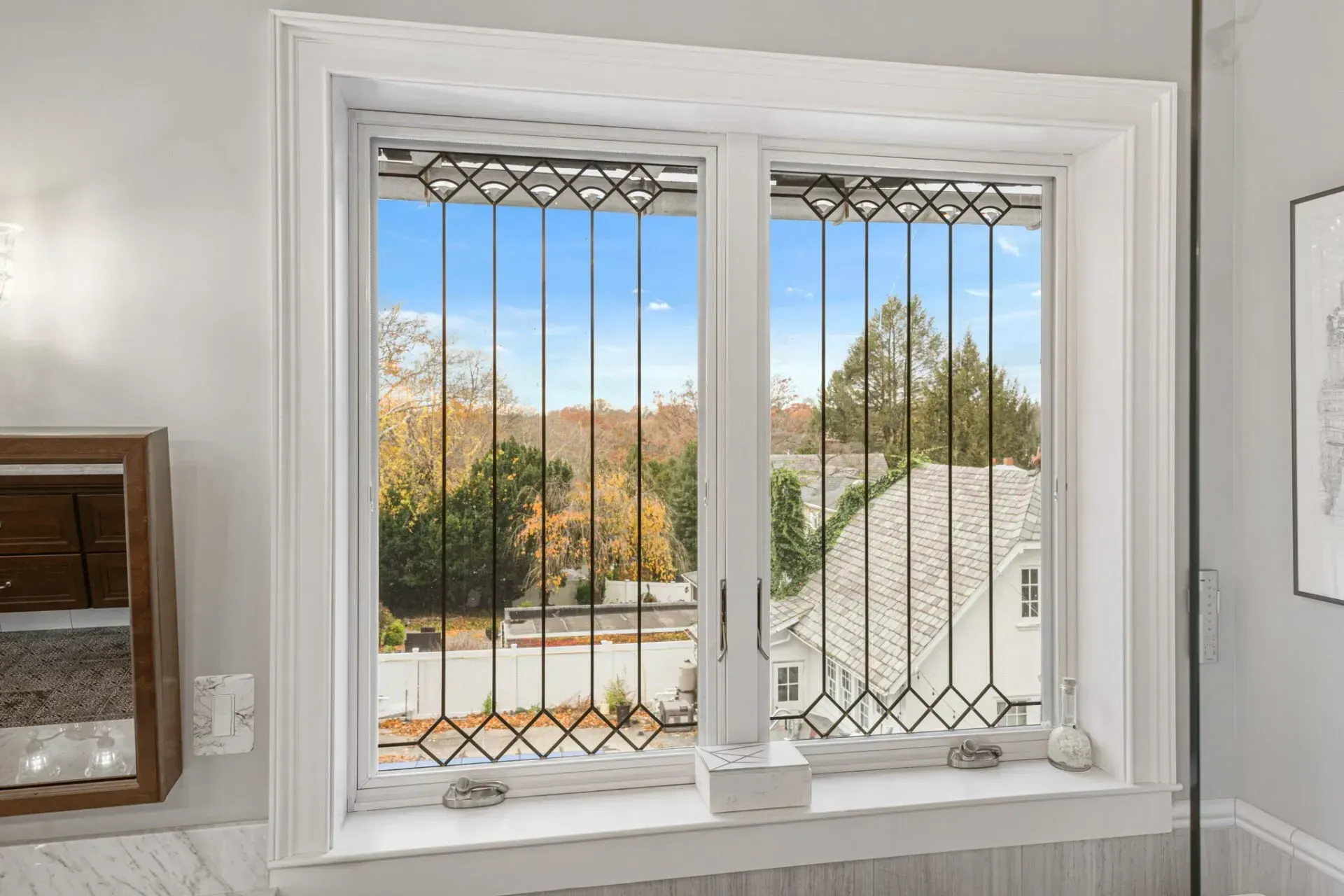 White-framed bay window with decorative metal grilles, overlooking autumn trees and a rooftop outside. in Bucks County PA