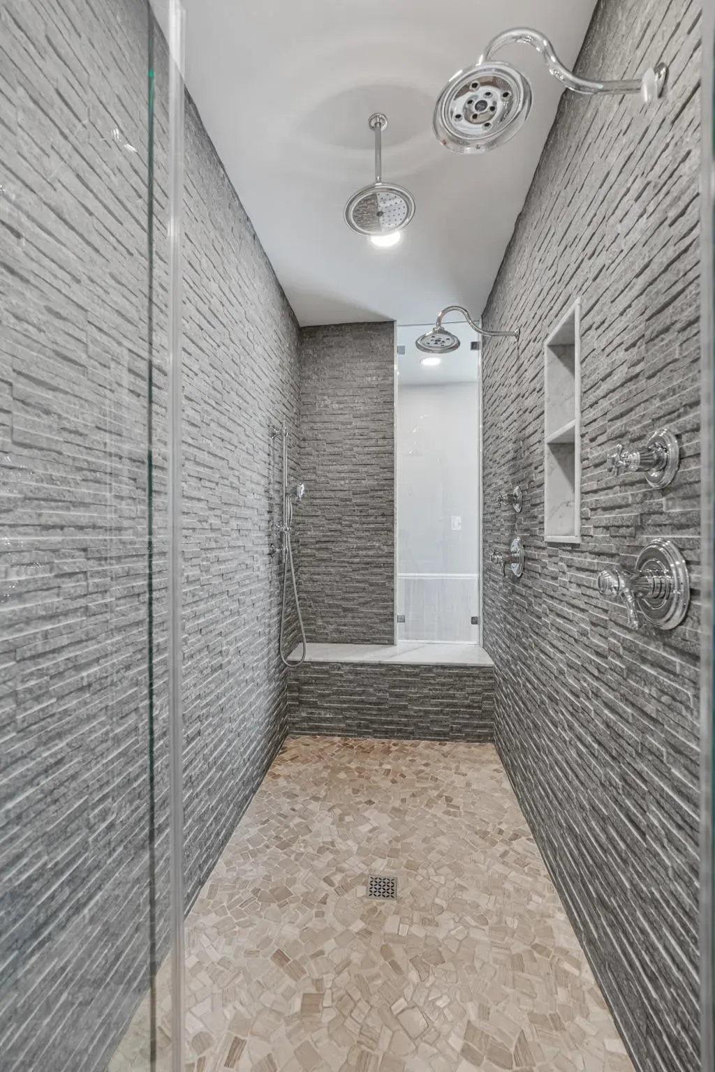 Narrow tiled shower corridor with gray brick walls, glass shelf, and white fixtures In Bucks Country PA