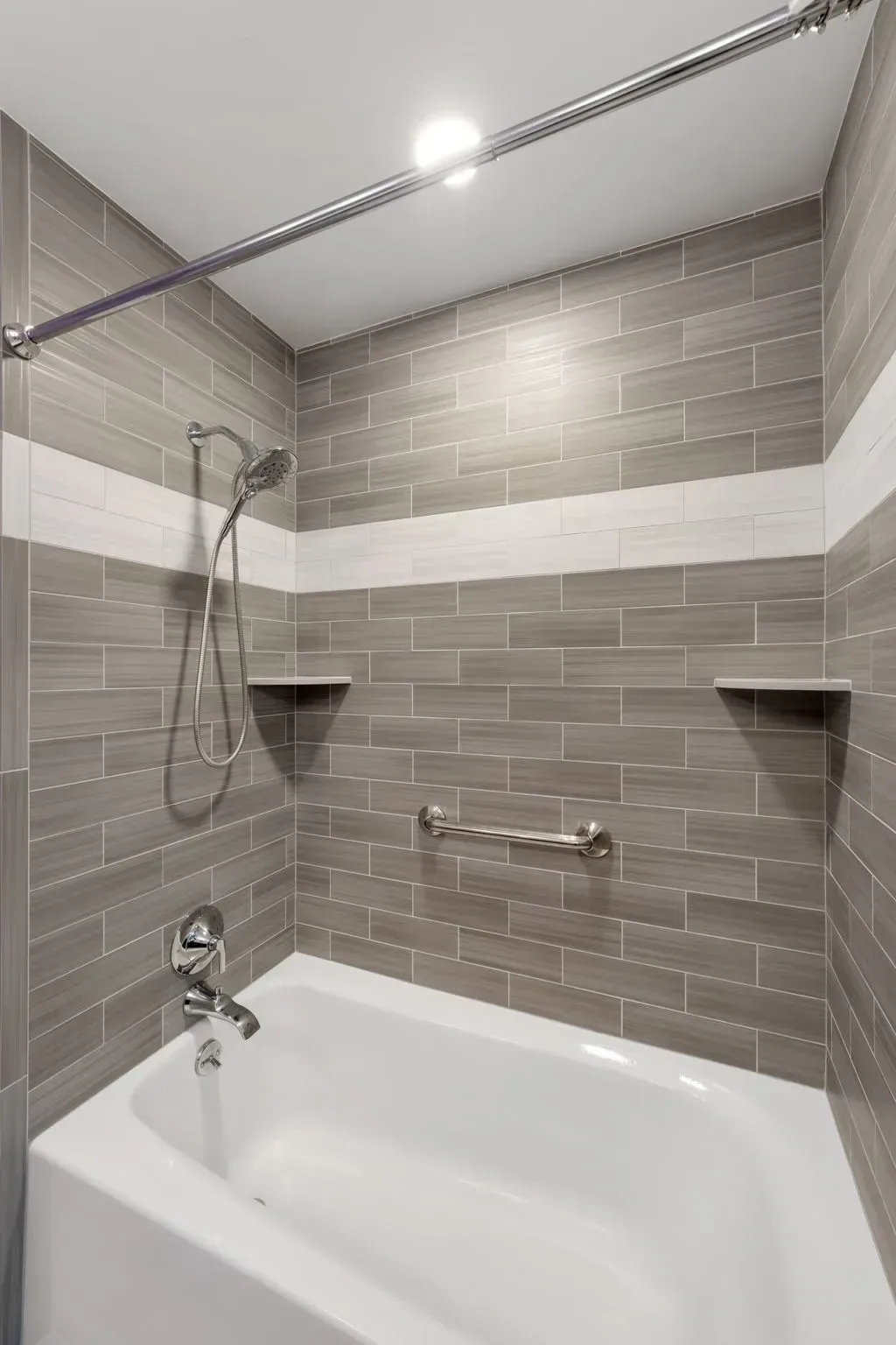 Small tiled bathroom with a shower-over-tub, gray walls, white accent stripe, and built-in corner shelves In Bucks County PA