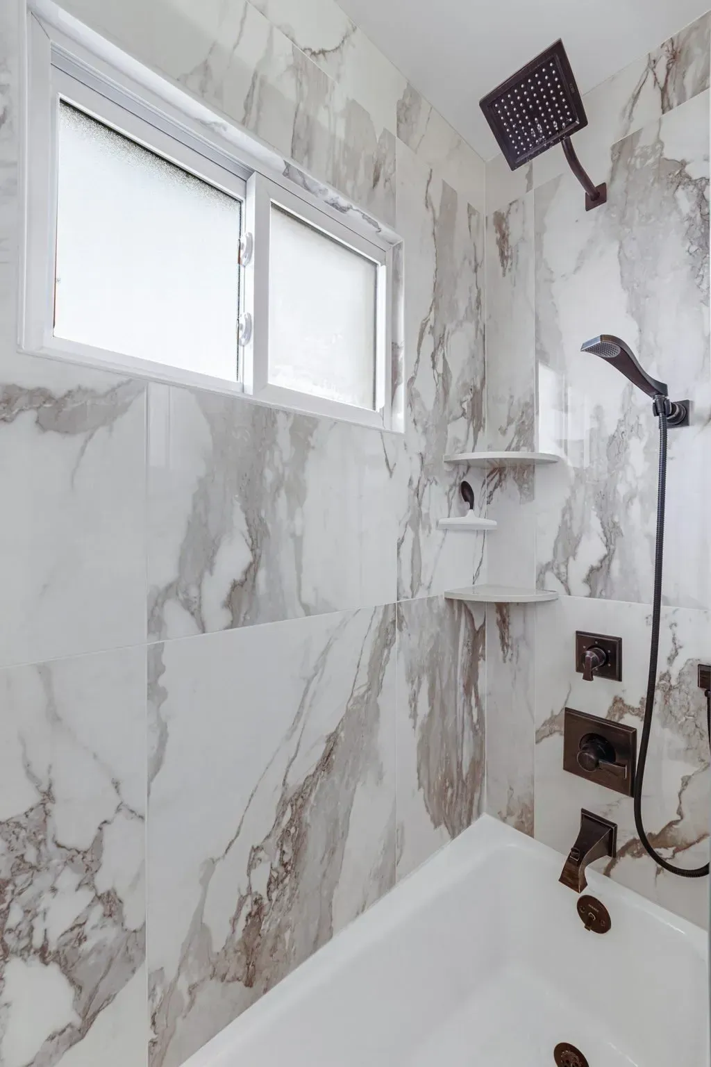 White tiled bathroom with a window, showerhead, shelf, and bathtub corner In Bucks County PA