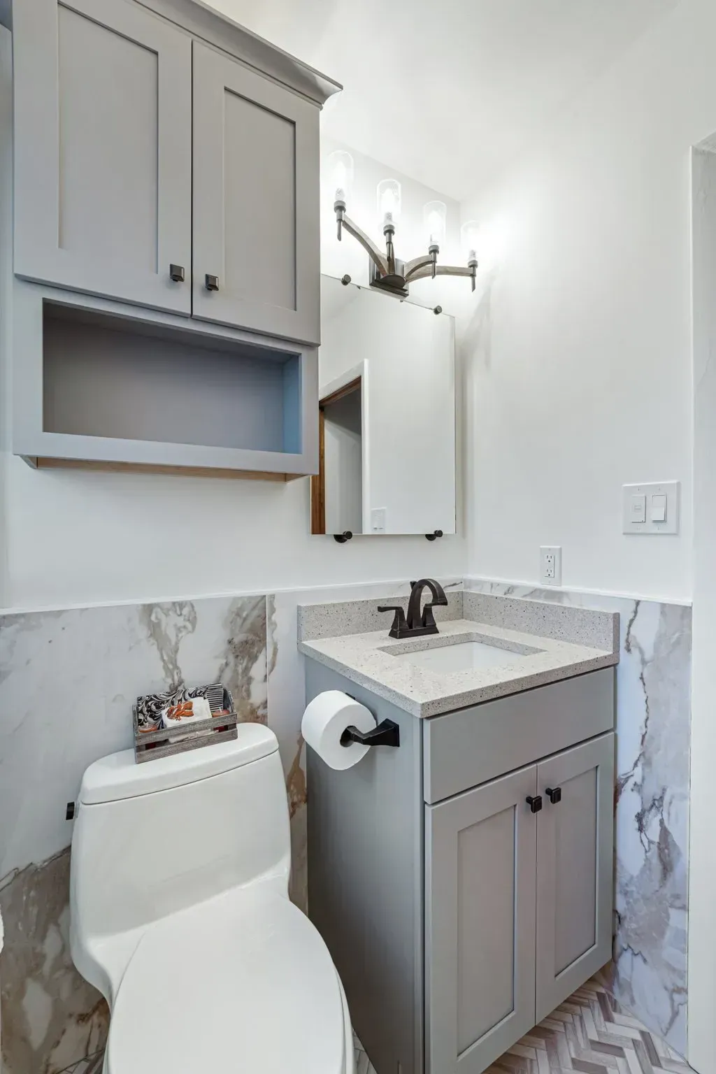 Small powder room with gray vanity, toilet, marble wall tile, and brass light fixture In Bucks County PA