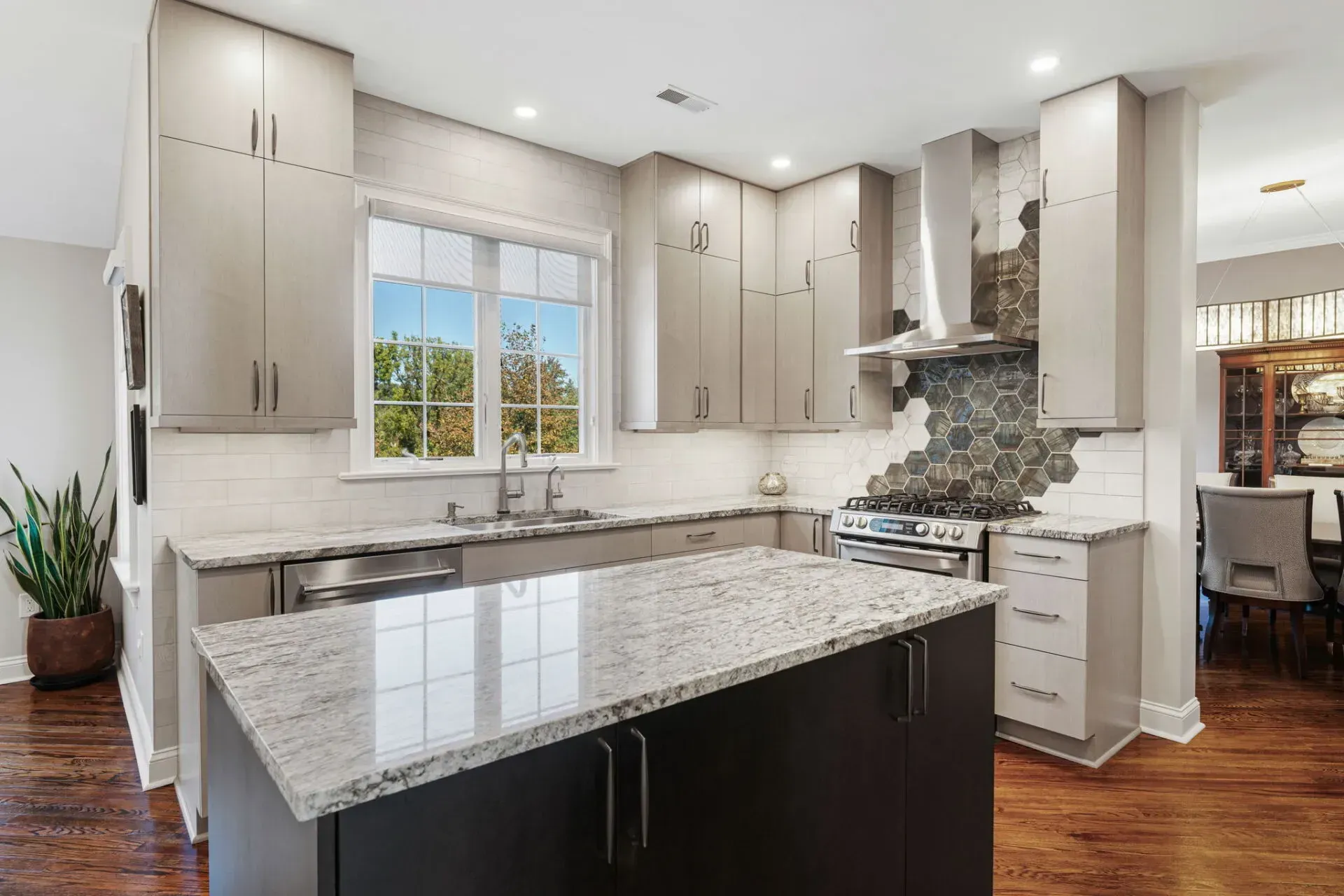 Modern white kitchen with granite island, stainless fixtures, and stone backsplash, lit by a window and recessed lights In Bucks County PA