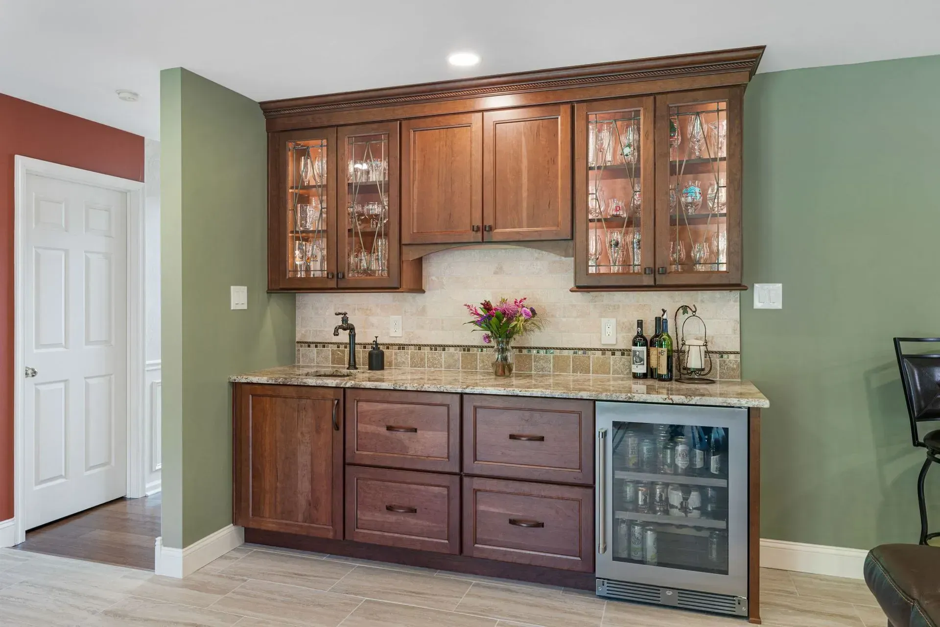 Kitchen bar with wooden cabinets, granite countertop, and glass-front drink fridge against green walls  In Bucks County PA