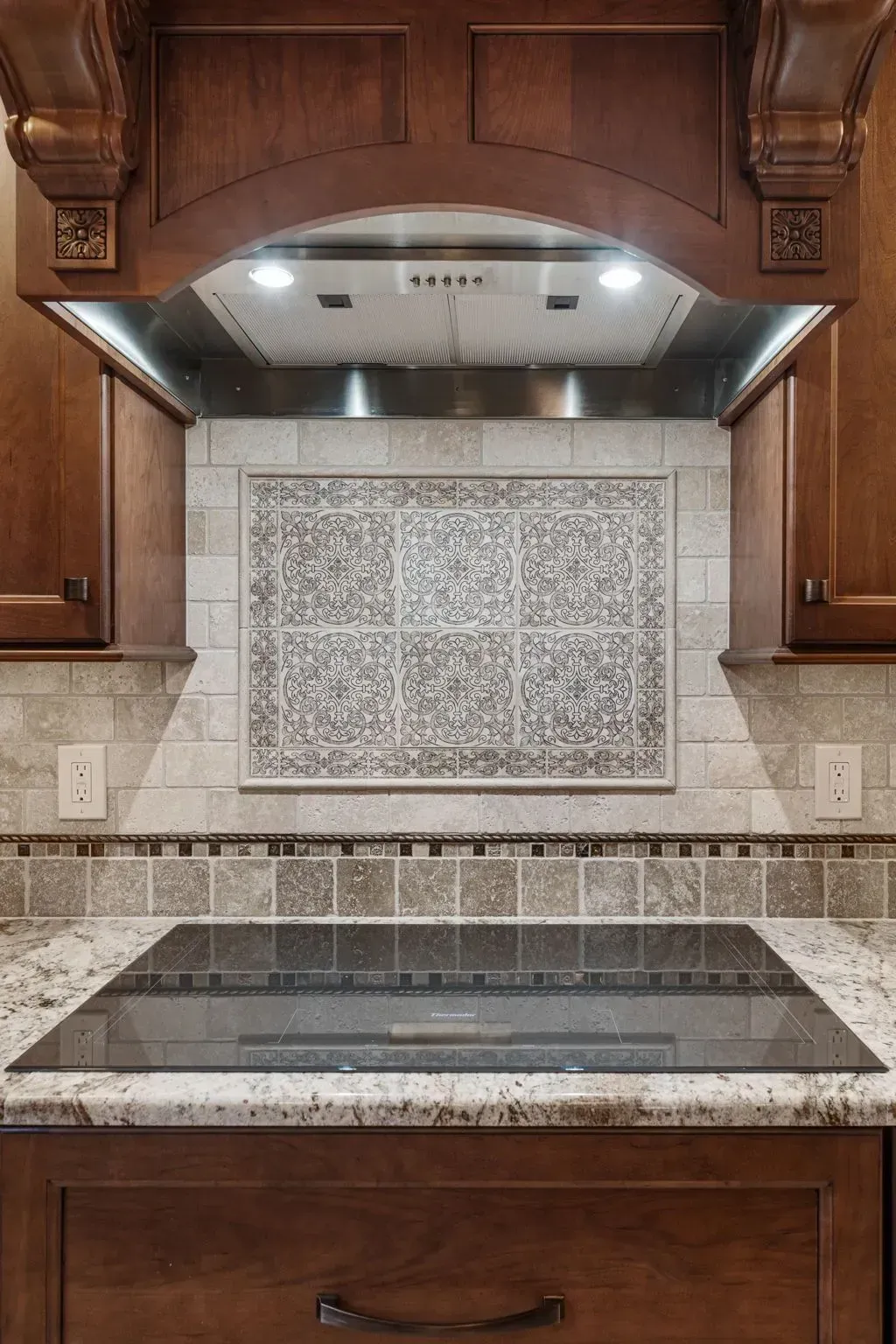 Kitchen cooktop with stainless hood, wooden cabinets, tiled backsplash, and granite countertop  In Bucks County PA