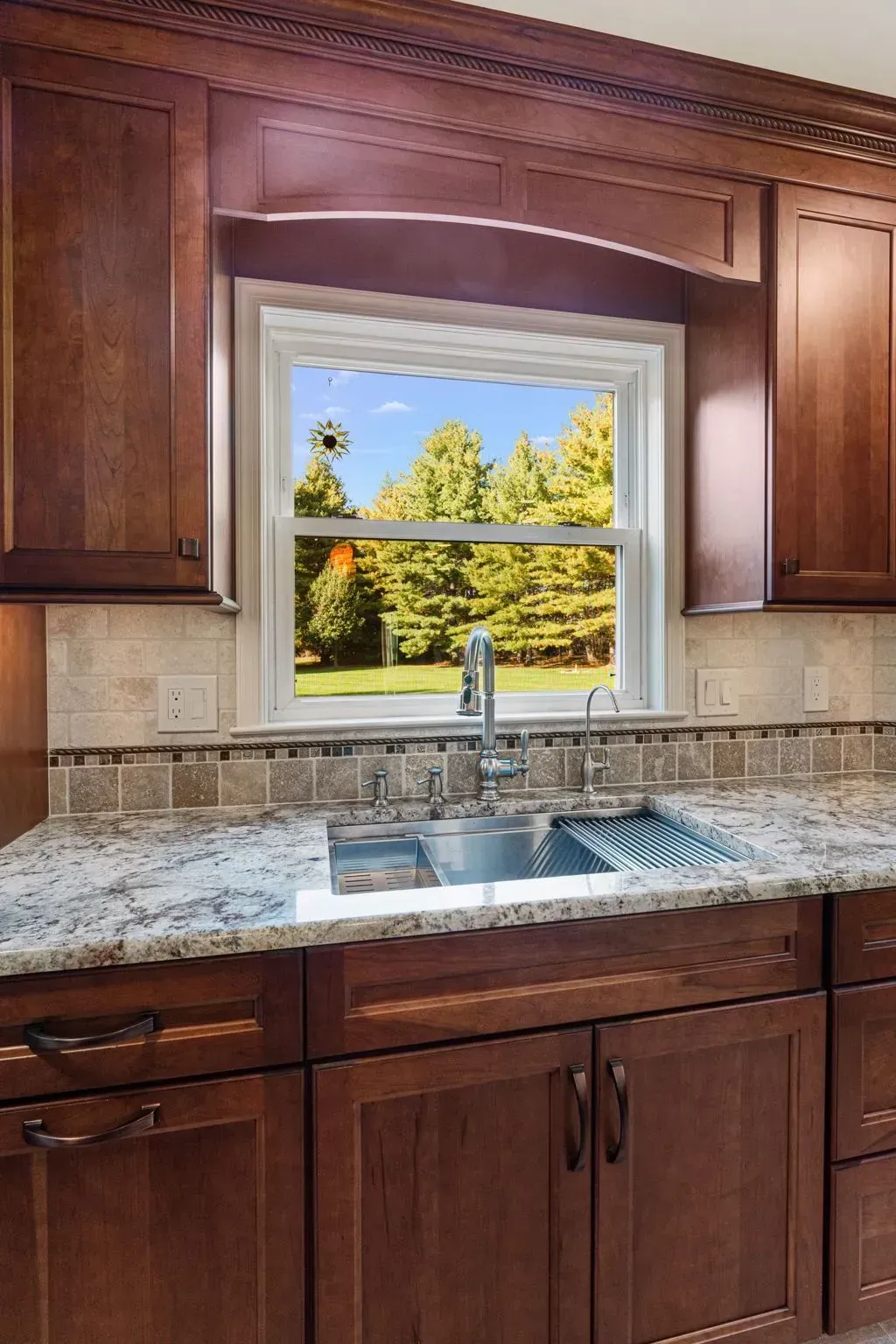 Kitchen sink beneath window, with dark wood cabinets and granite countertop overlooking trees.