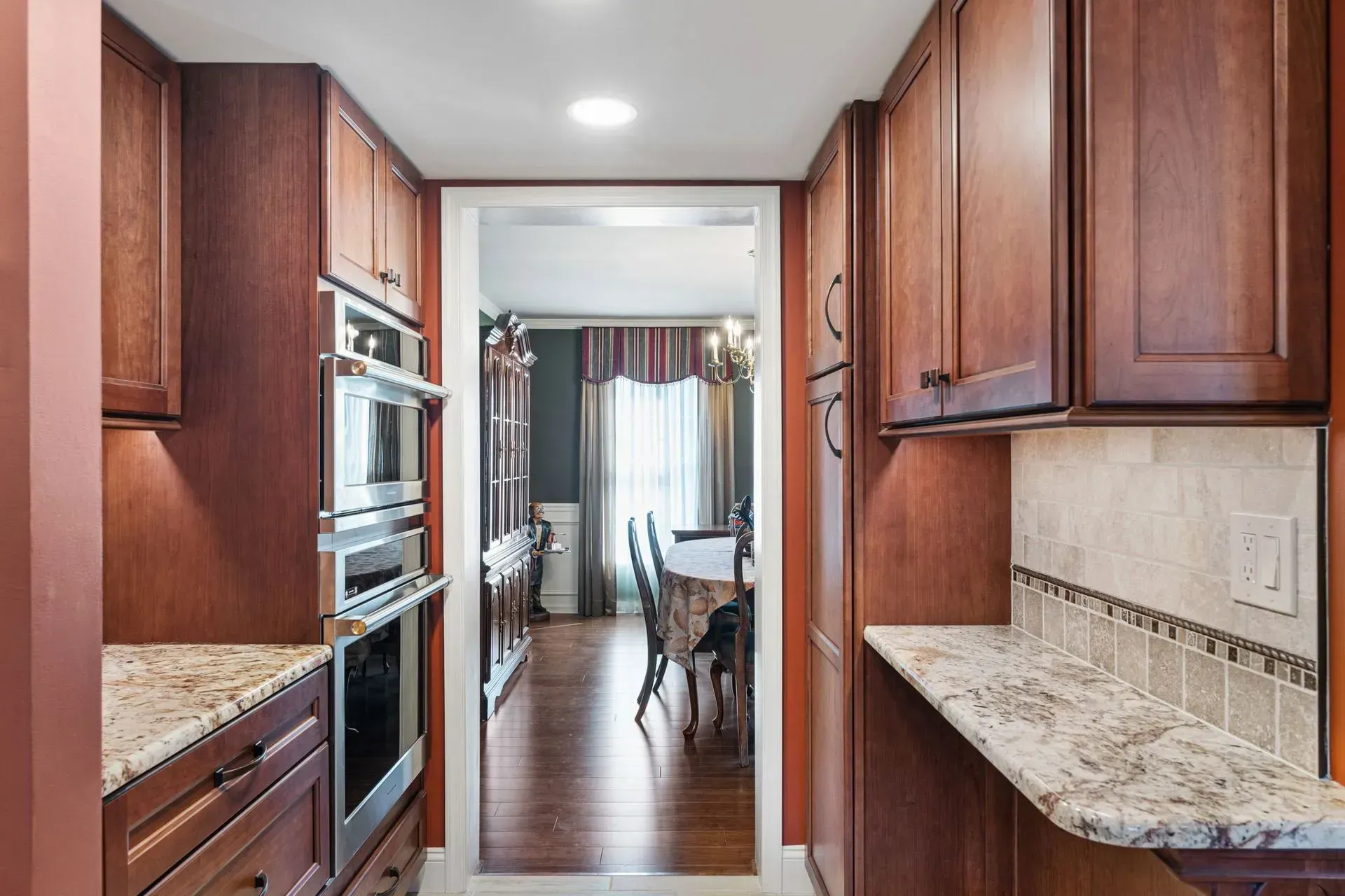 Narrow kitchen with cherry cabinets, granite counters, and a view into a dining area with curtains  In Bucks County PA