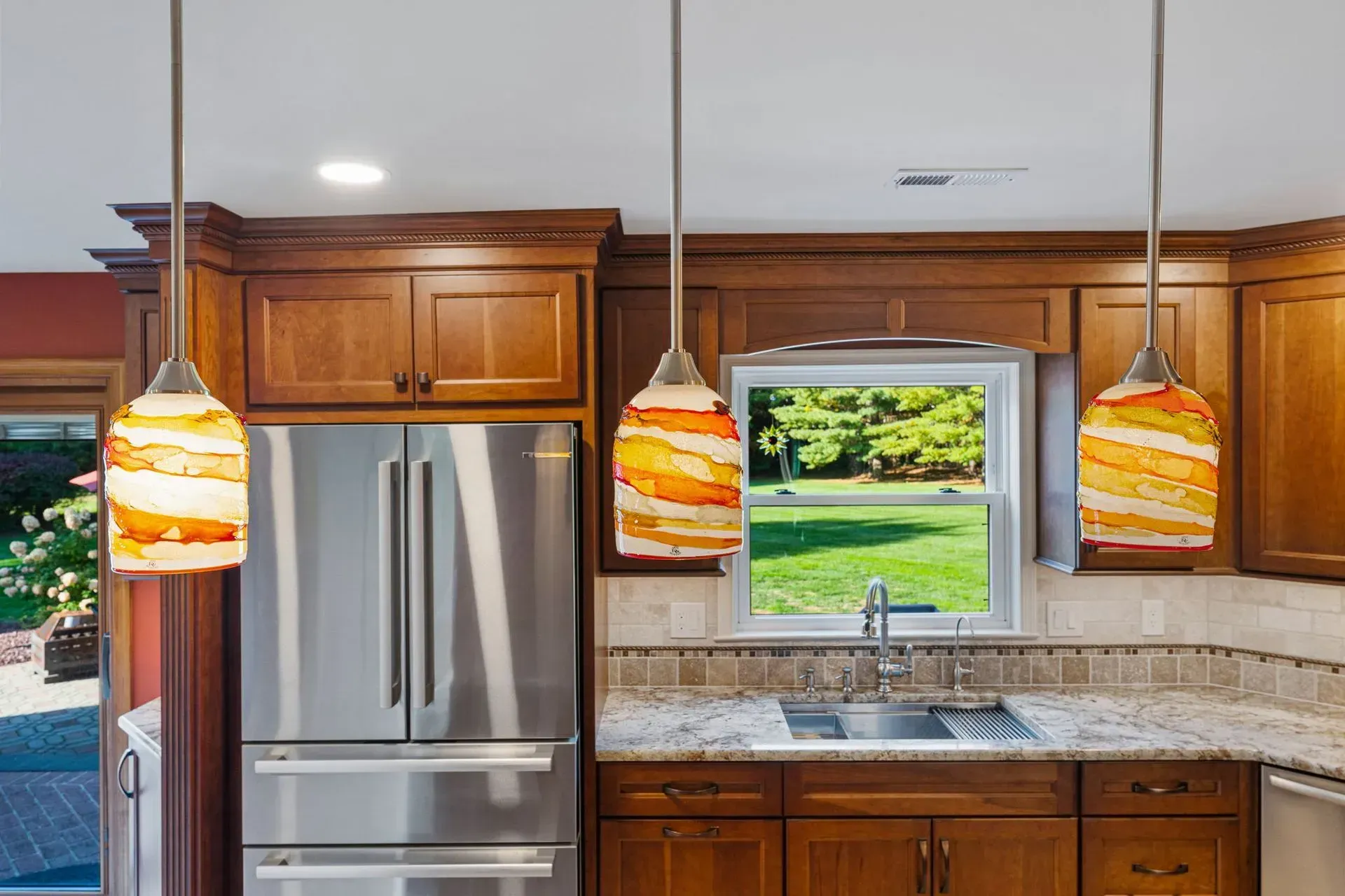Modern kitchen with stainless fridge, wooden cabinets, granite counter, and three pendant lights over a sink window  In Bucks County PA