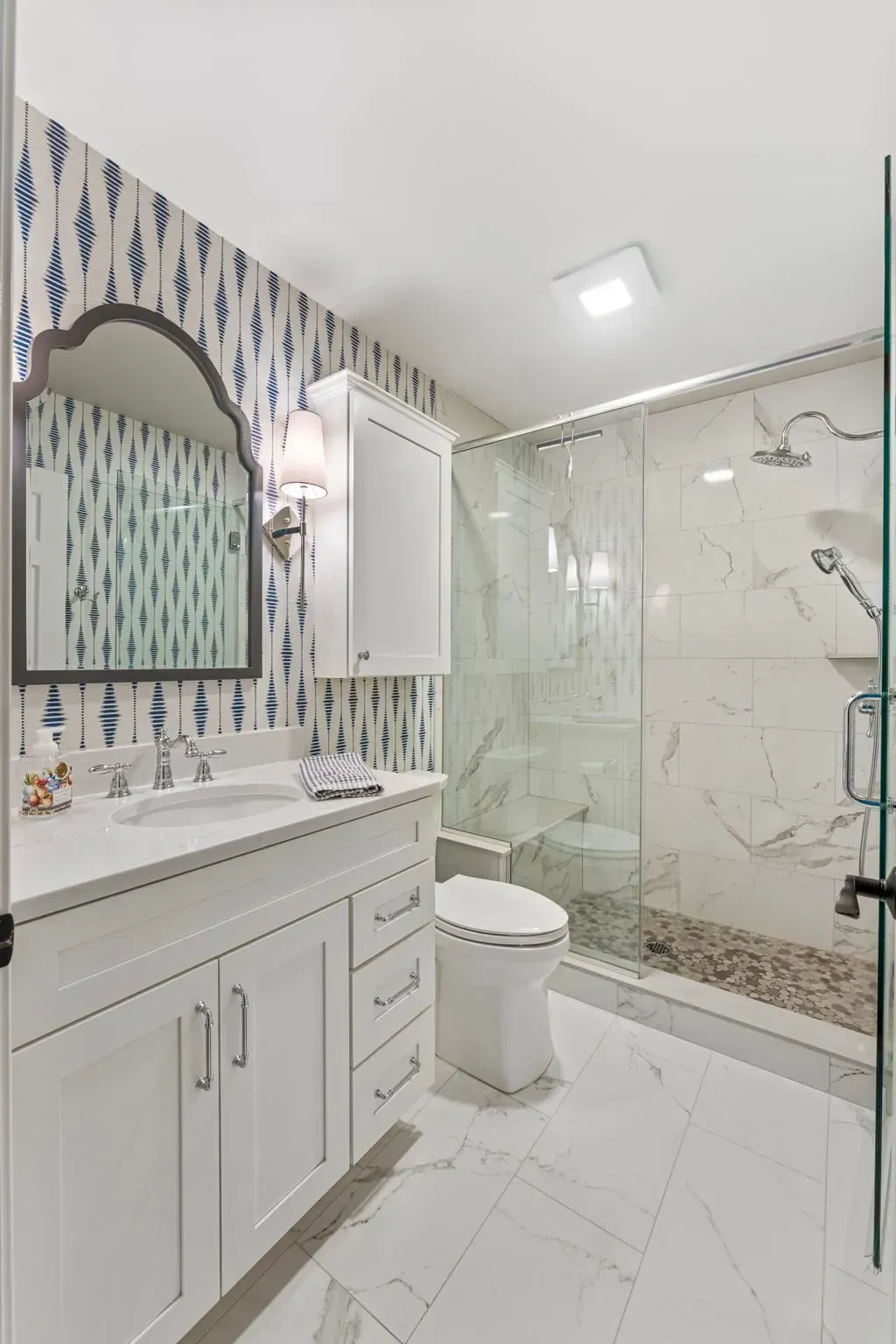 Bright bathroom with white vanity, patterned wallpaper, mirror, toilet, and glass shower enclosure  In Bucks County PA