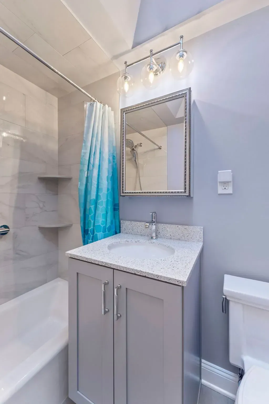 Small bathroom vanity with mirror, light gray cabinets, white countertop, and blue shower curtain beside a tub  In Bucks County PA
