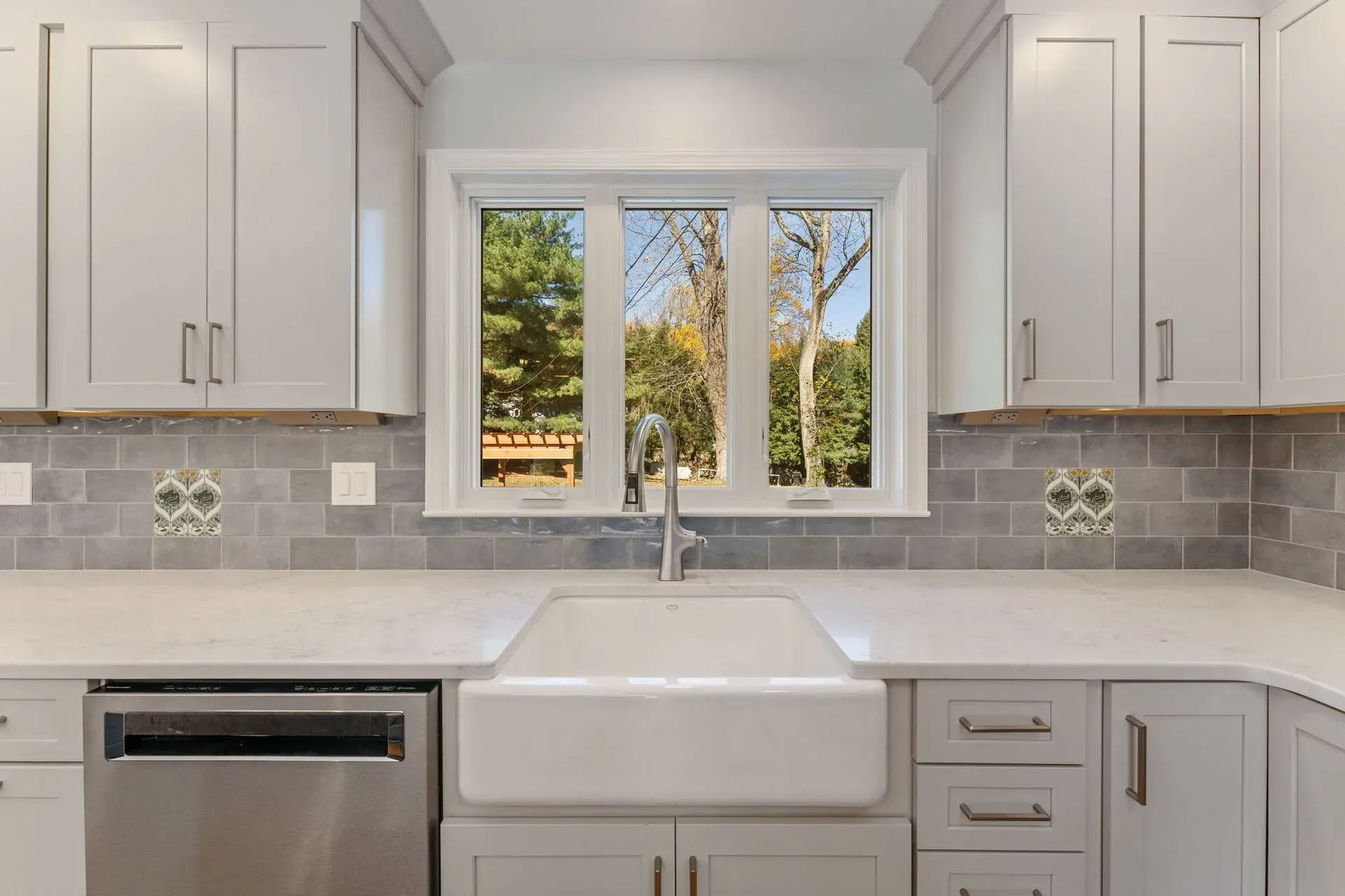 Bright white kitchen with farmhouse sink beneath a window, gray backsplash, and white cabinets  In Bucks County PA