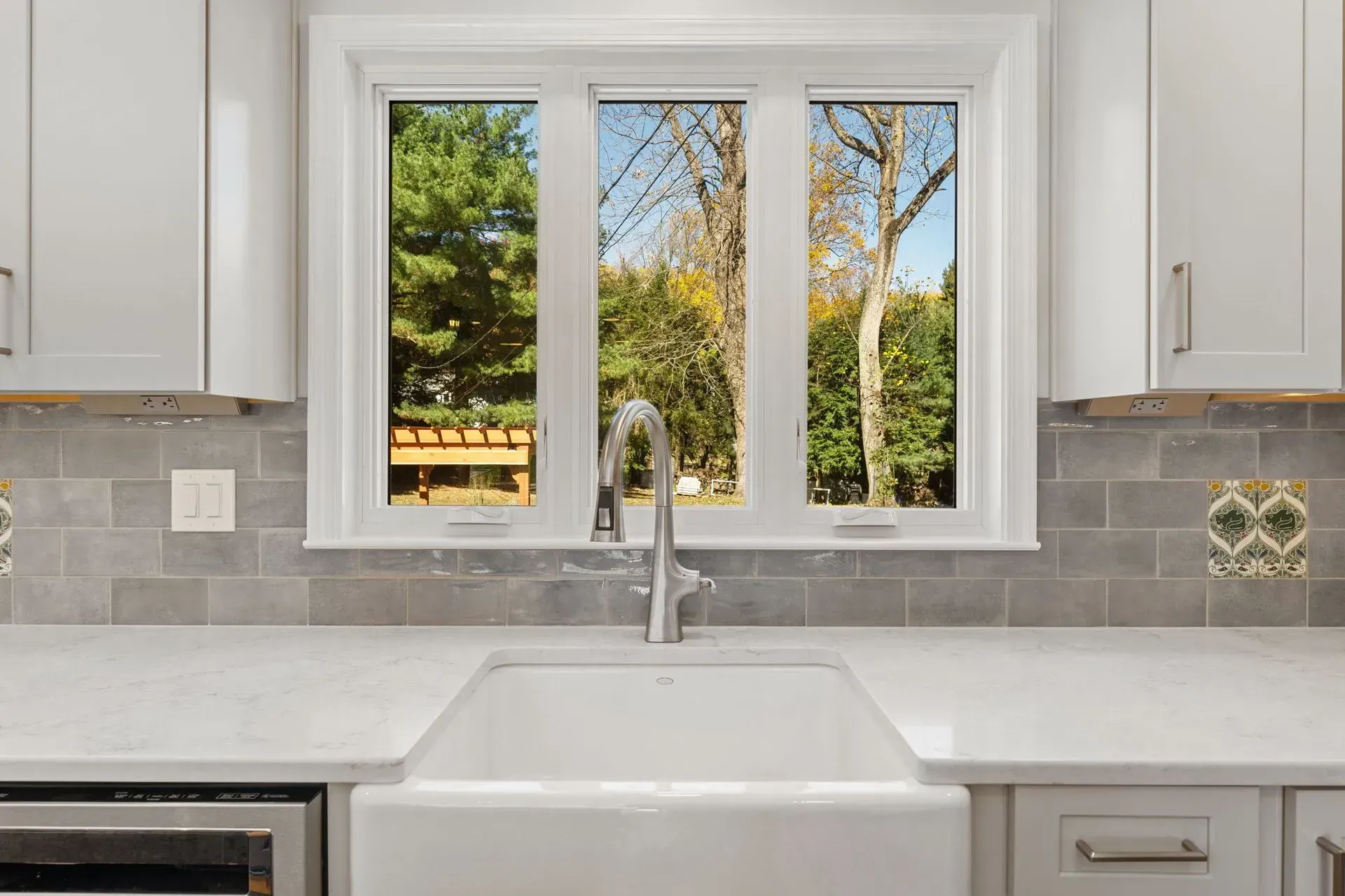 Bright kitchen sink beneath window, with white cabinets, gray tile backsplash, and garden view outdoors  In Bucks County PA