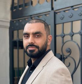 A man with a beard wearing a tan jacket and black shirt