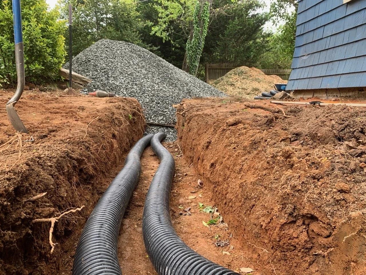 Trenches with black drainage pipes leading to a gravel pile beside a blue house