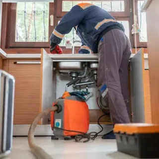 Plumber in blue and orange workwear, working on kitchen sink plumbing with an orange pump.