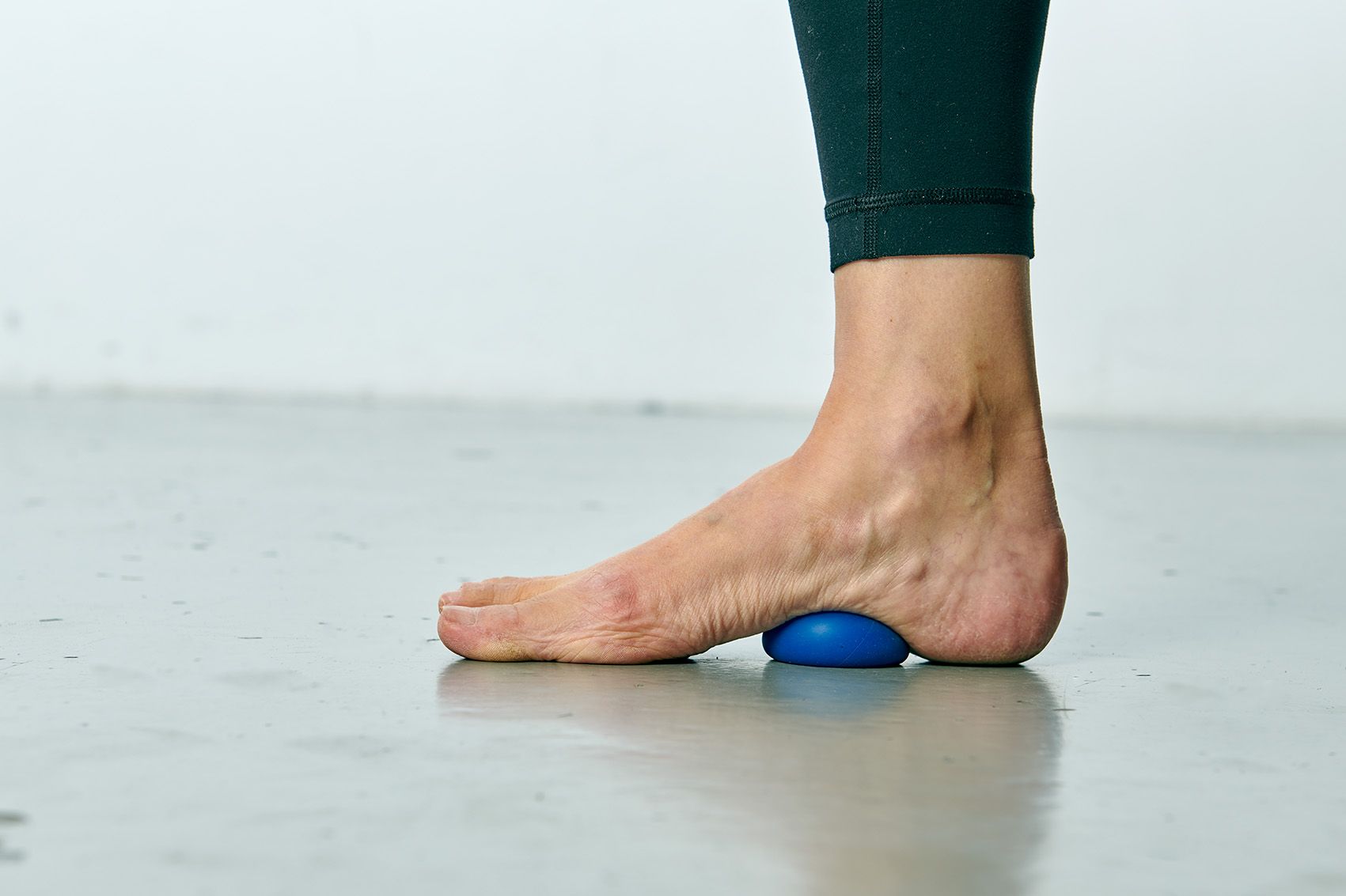 Fussmassage eines blauen Balls auf grauem Boden. Die Person trägt eine dunkelgrüne Hose.