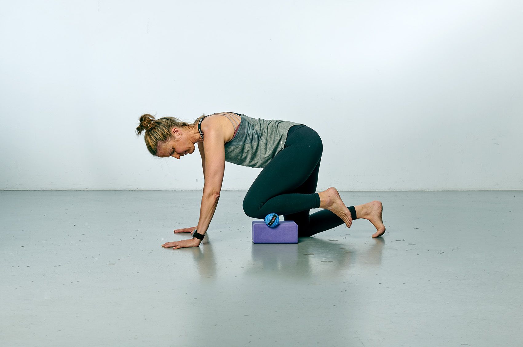 Frau in Yoga-Pose, Hände auf dem Boden, Knie auf einem Block, Blick nach unten. Barfuss, in einem Innenraum.