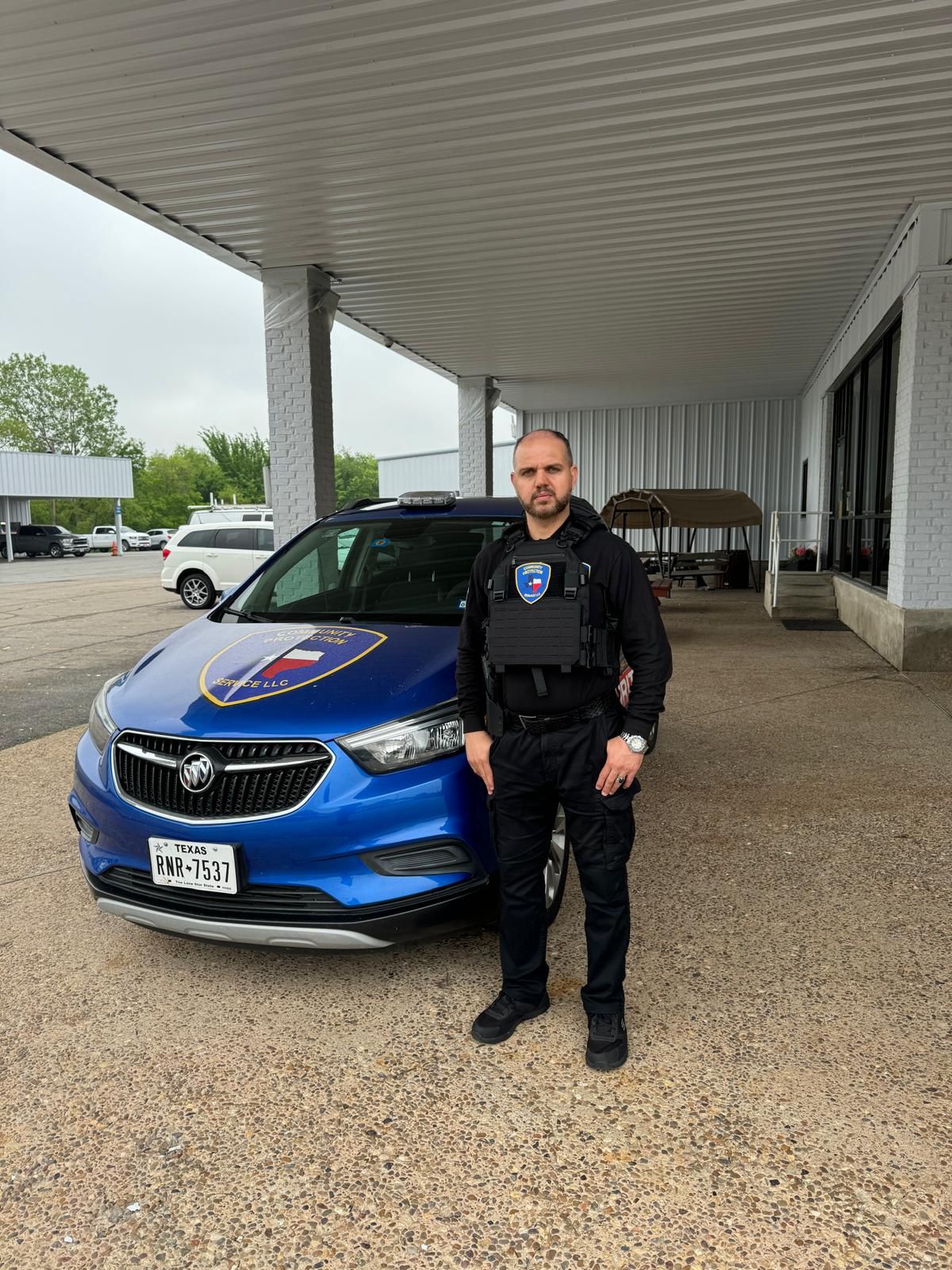 Man in black uniform standing beside a blue SUV under a covered driveway