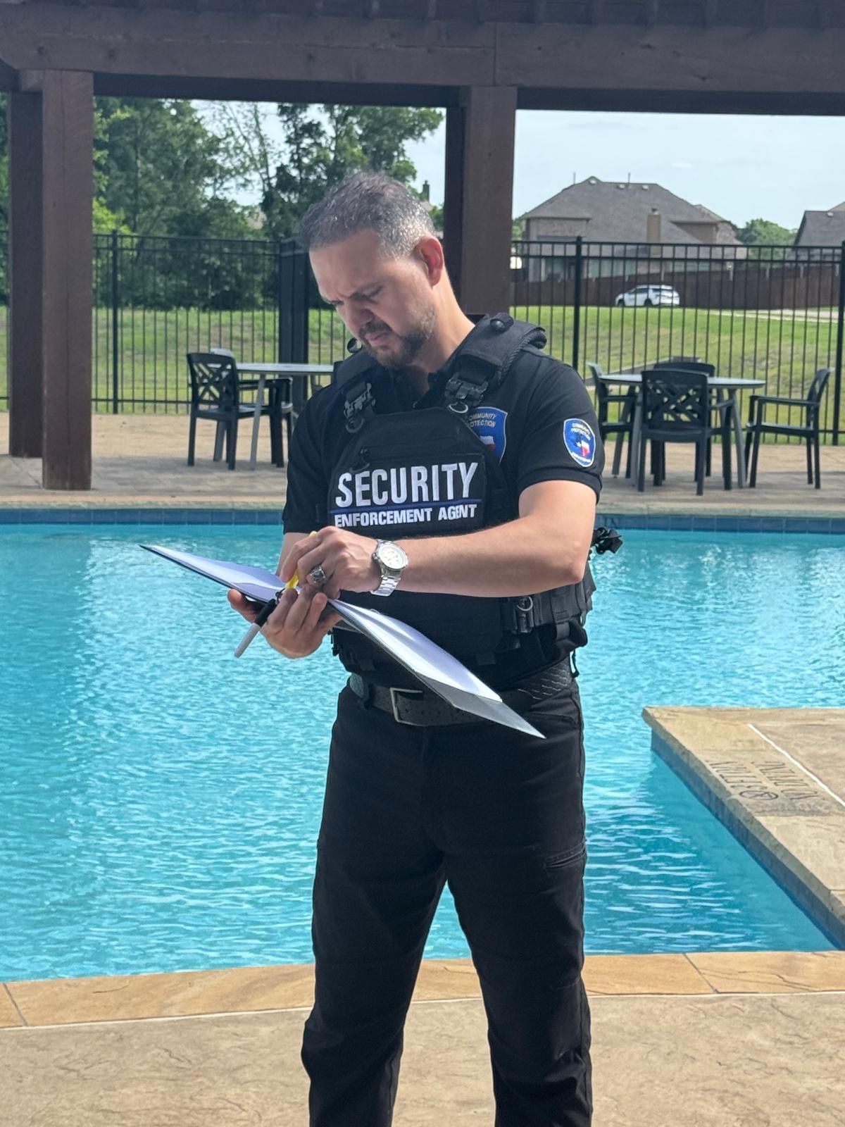 Security guard by a pool reading a clipboard, with chairs and a fence in the background.