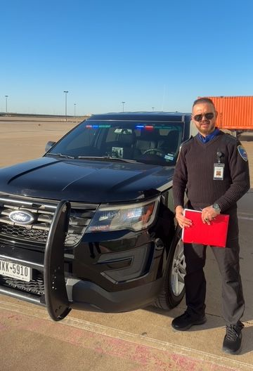 Man in dark jacket stands beside a black police SUV holding a red folder in a sunny parking lot.