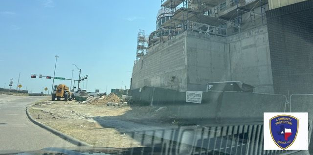 Roadside construction site beside a large concrete building, with heavy equipment and an unfinished overpass.