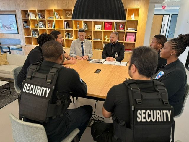 Security officers meet with two men around a conference table in a bright office.