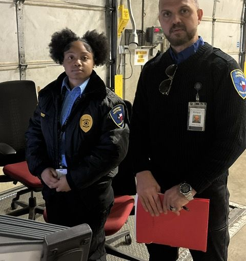 Two uniformed security officers stand in an office, one holding a red folder.