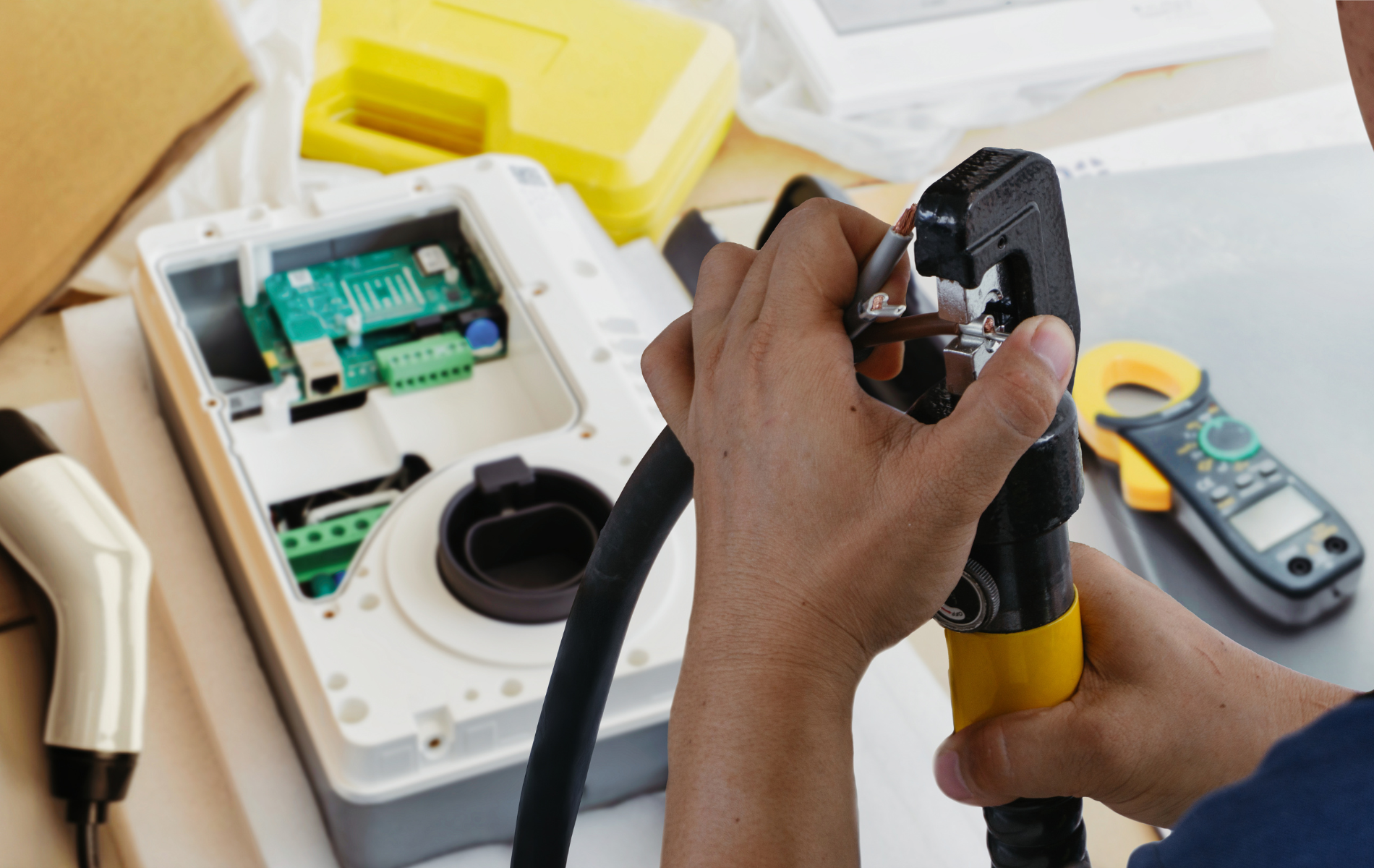 Person using a crimping tool on an EV charger cable. Electronics and tools are visible on a table.