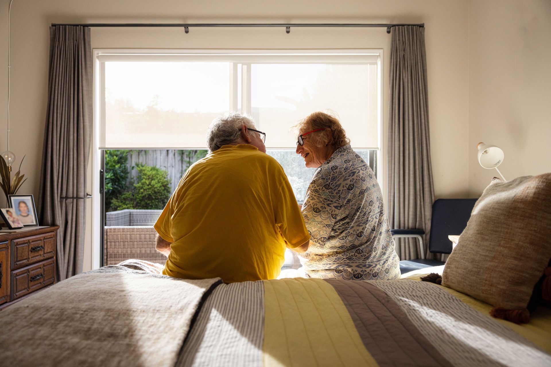 An elderly couple is sitting on a bed looking out a window.