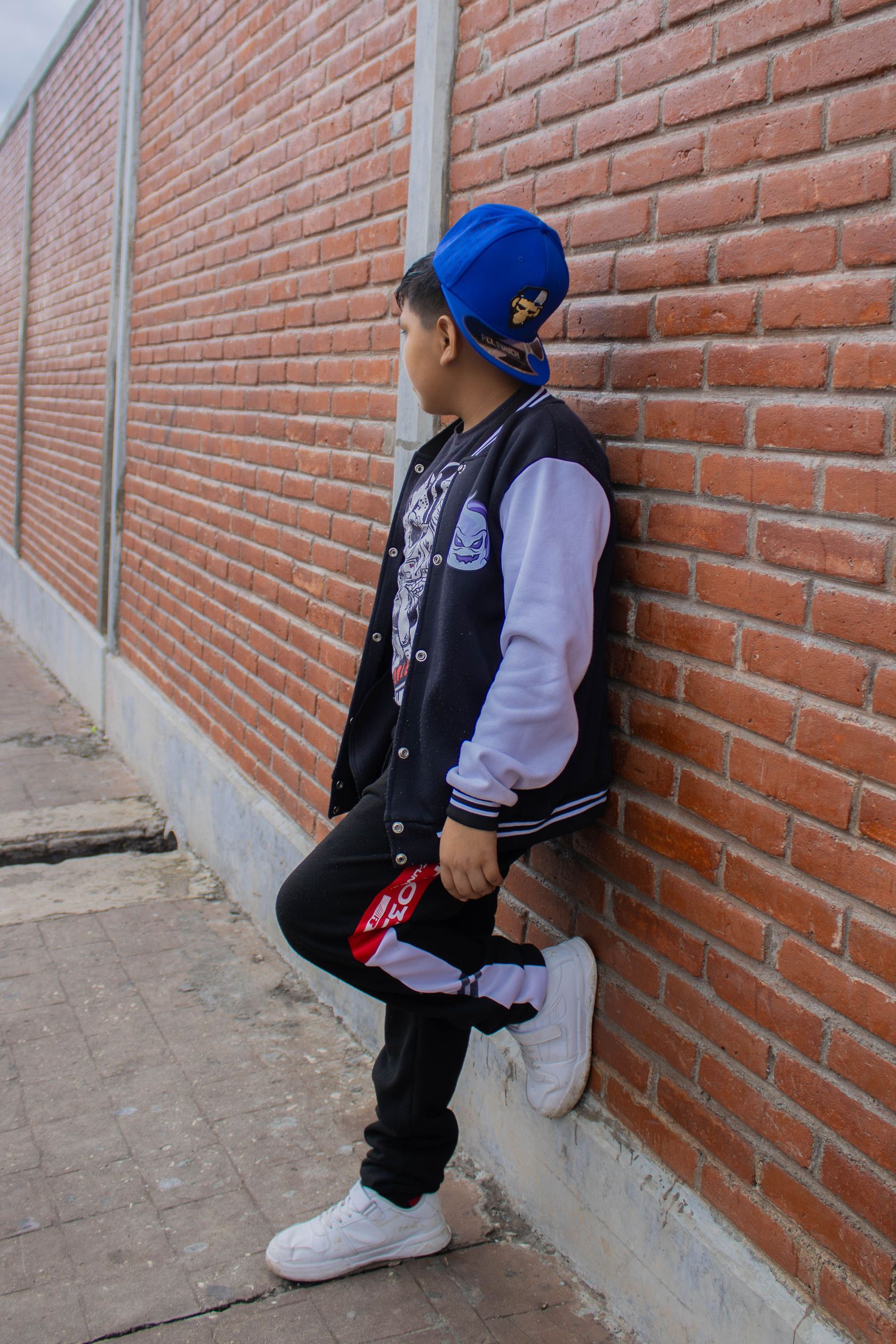 Persona apoyada contra una pared de ladrillos, con gorra azul, chaleco negro y zapatillas blancas.
