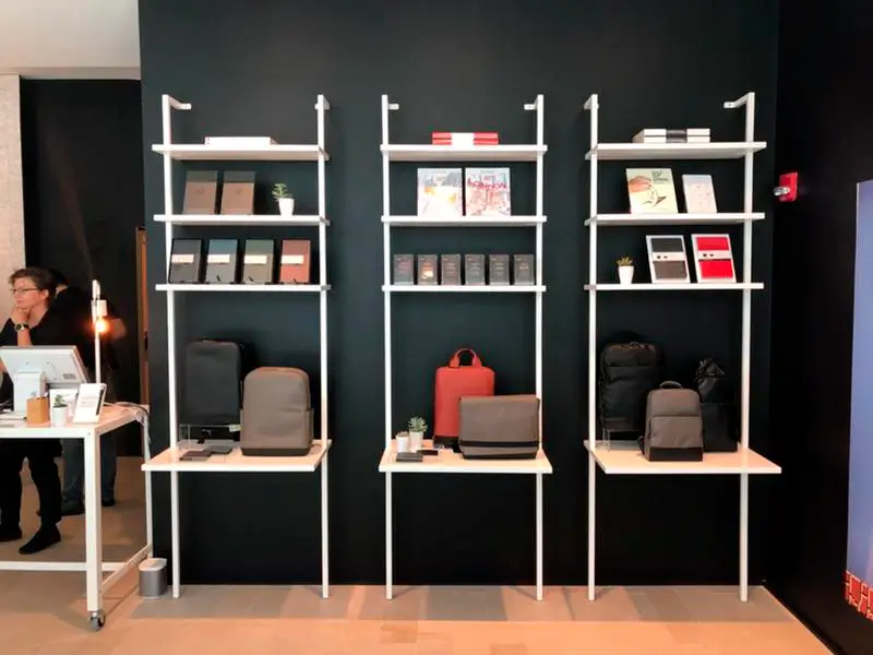 A woman sits at a desk in front of a wall with shelves filled with books and bags