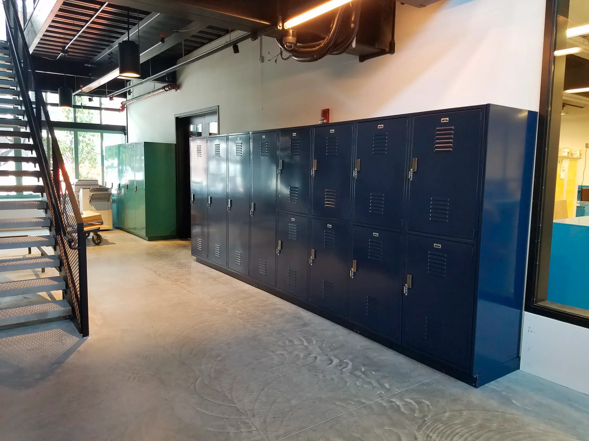 A row of blue lockers are lined up in a room next to stairs.