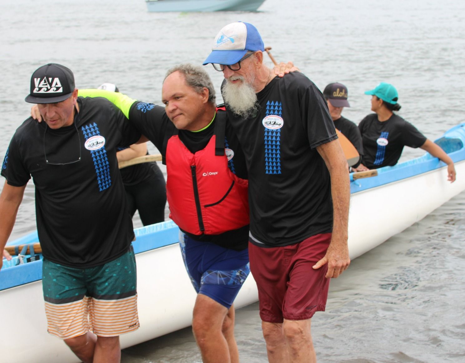 People carrying a white canoe through shallow water, one in a red life vest, others in black shirts and caps