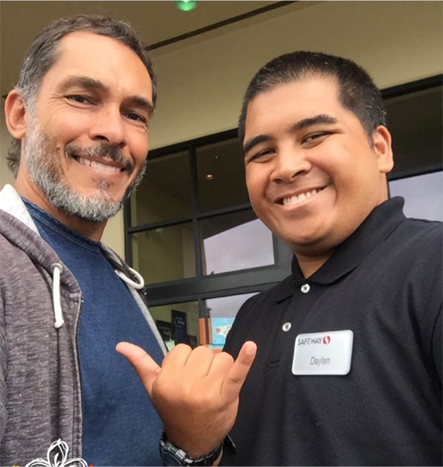 Two smiling men posing outdoors, one making a shaka hand gesture, near a building entrance.