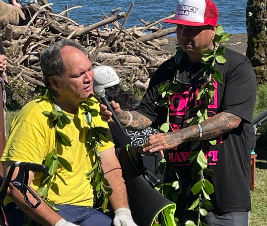 A man in a yellow shirt is talking into a microphone while another man in a red hat looks on.